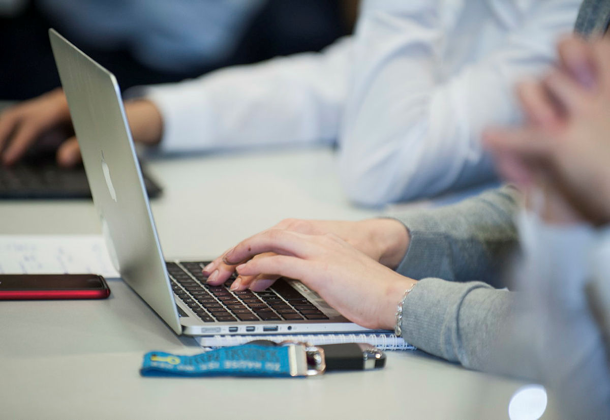 Person tippt auf Apple-MacBook in einer Besprechung mit Smartphone und USB-Stick auf dem Tisch.